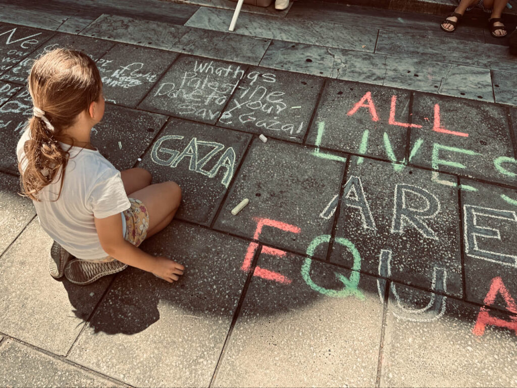 A young girl kneels among chalk writing. She has just written the single word GAZA. Alongside, in the colours of the Palestinian flag is written 'All lives are equal' in pavement chalk.