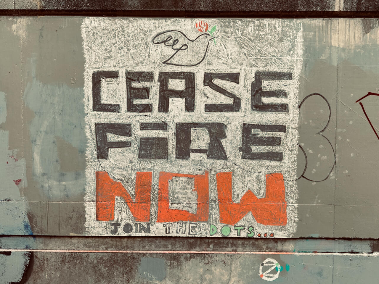 Chalk mural on a wall. A dove with red and green roses above the words 'Ceasefire Now' and the phrase 'Join the dots...' Small dotZero symbol below. dotZero symbol.