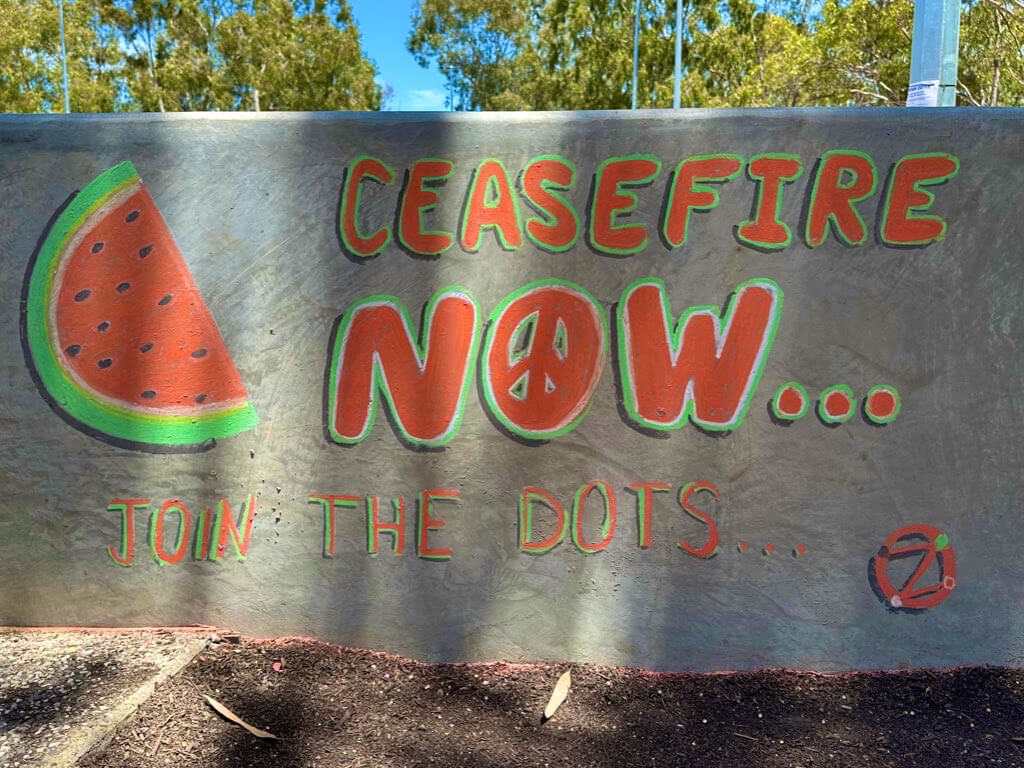 The phrase 'Ceasefire Now'chalked on a wall in bold Palestinian colours with a half watermelon alongside. The phrase 'Join the dots...' underneath. DotZero symbol too.