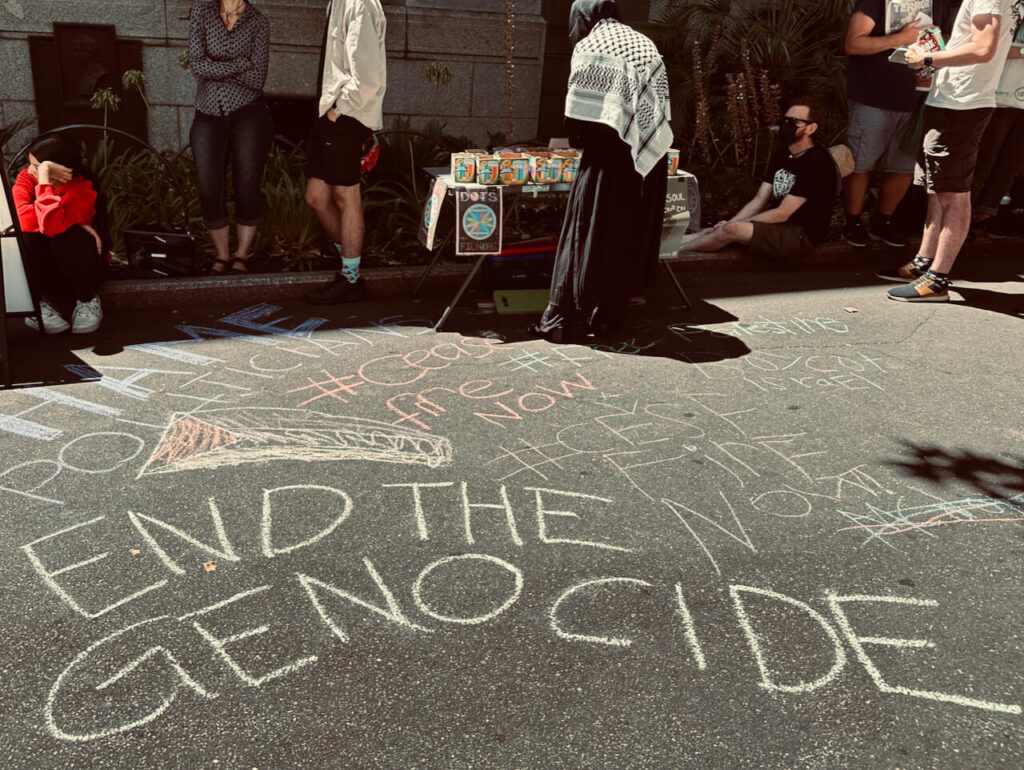 'End the genocide', Palestinian flag, hashtag #Ceasefirenow written in pavement chalk. A woman in Palestinian dress is selecting chalk from a nearby table with DOTS poster.