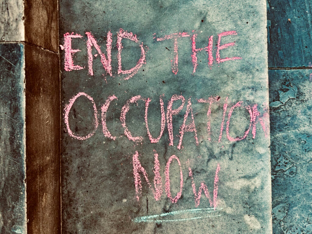 'End the occupation now' scrawled in pavement chalk on wall. All caps writing. The word 'NOW' underlined for emphasis.