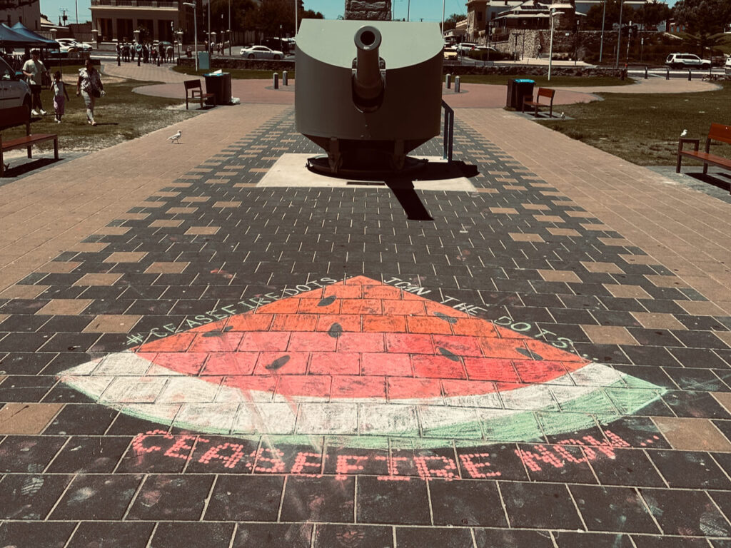 Large watermelon in pavement chalk with 'ceasefire now' written in the foreground. Also the hashtag #ceasefiredots and phrase 'join the dots...'. WW1 naval cannon behind.